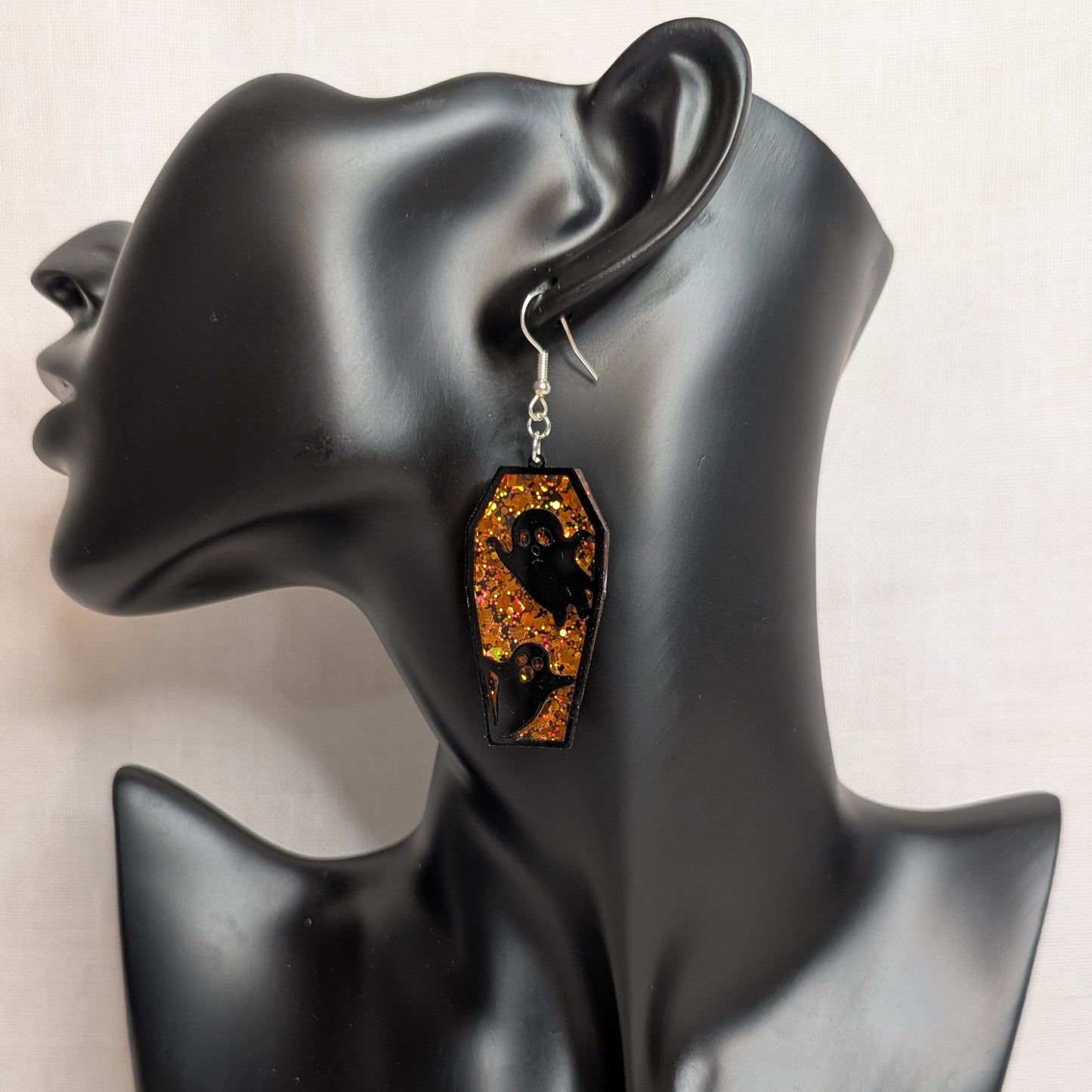 Orange glitter coffin-shaped dangle earring with black ghost cutouts displayed on a black jewelry mannequin