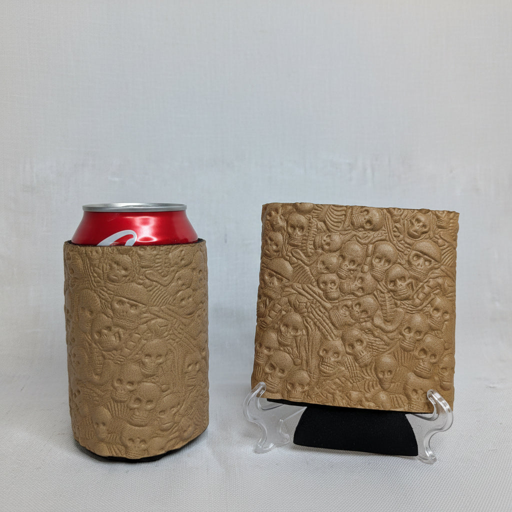 Tan embossed skull pattern leather can cooler holding a red beverage can beside a matching embossed leather sample on a display stand.