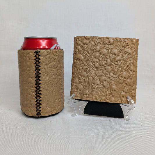 Tan embossed skull pattern leather can cooler with visible side stitching, red beverage can, and matching leather sample on a display stand.