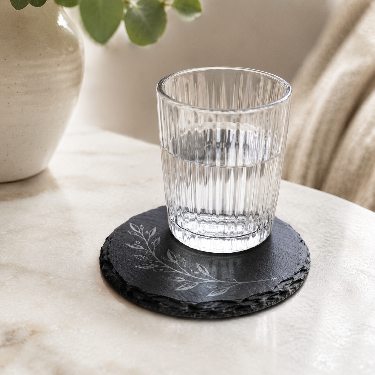 Clear drinking glass resting on a round engraved slate coaster with a light gray botanical design.
