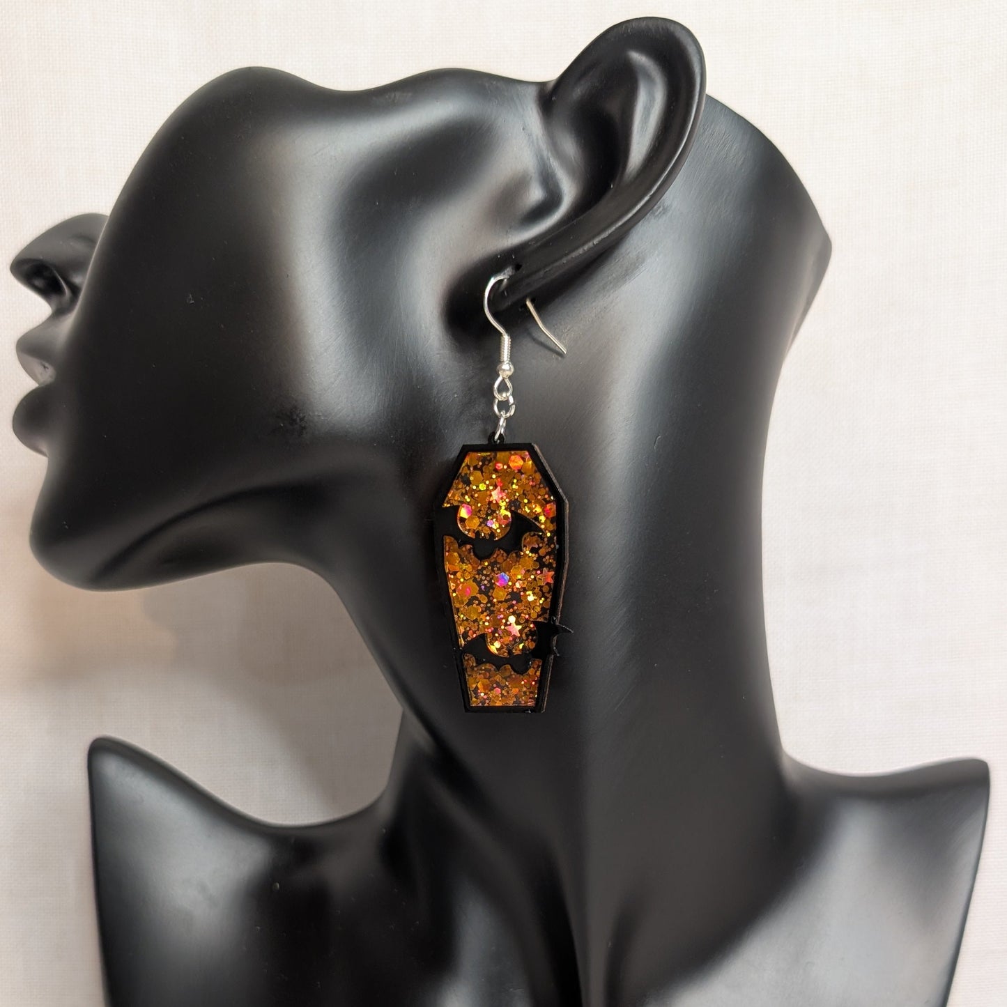 Orange glitter coffin dangle earring with black bat cutouts displayed on a black jewelry mannequin for Halloween.
