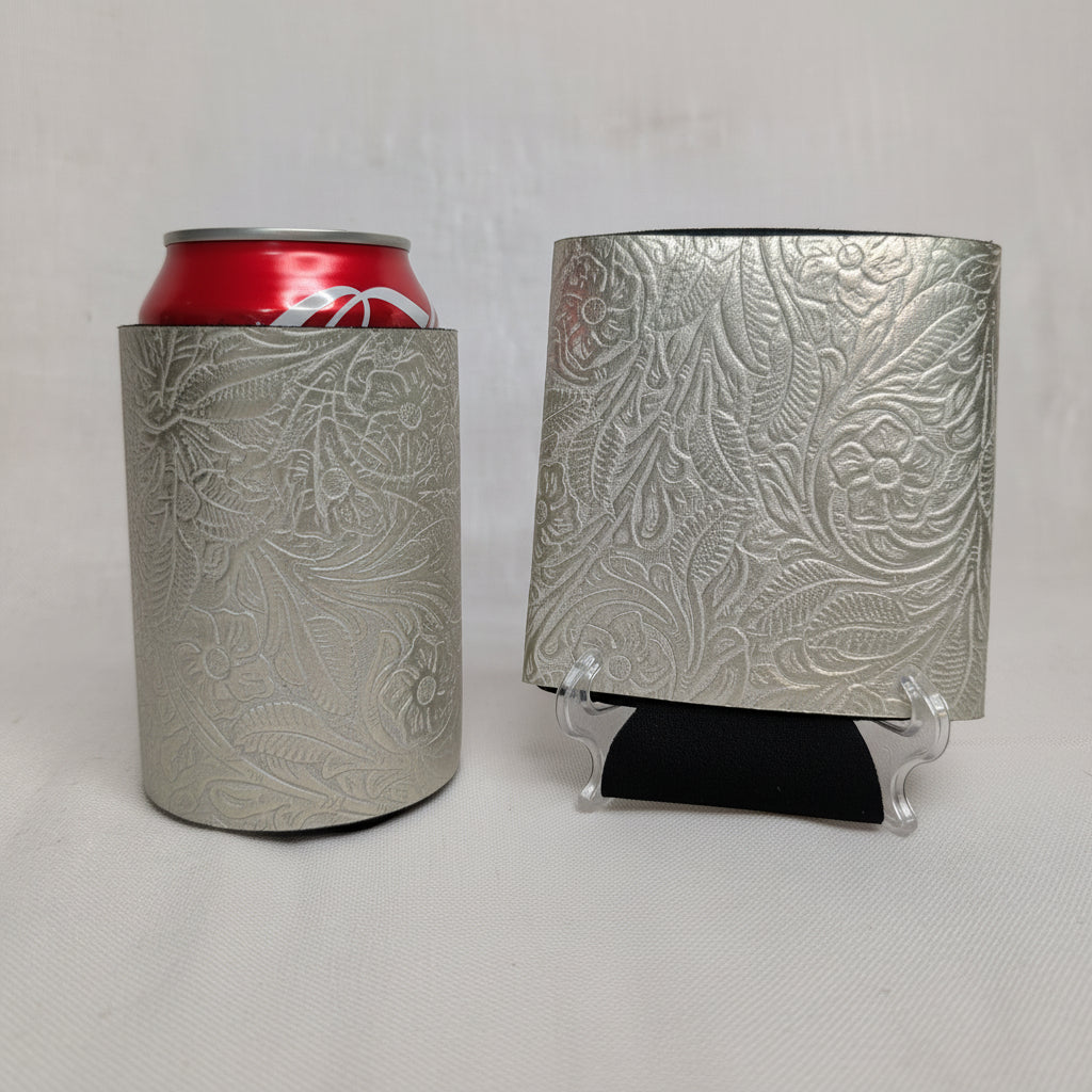 Silver metallic embossed floral leather can cooler holding a red beverage can beside a matching embossed leather sample on a display stand.