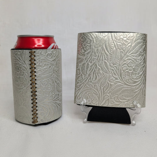 Silver metallic embossed floral leather can cooler with visible side stitching, a red beverage can, and matching embossed leather sample on a display stand.