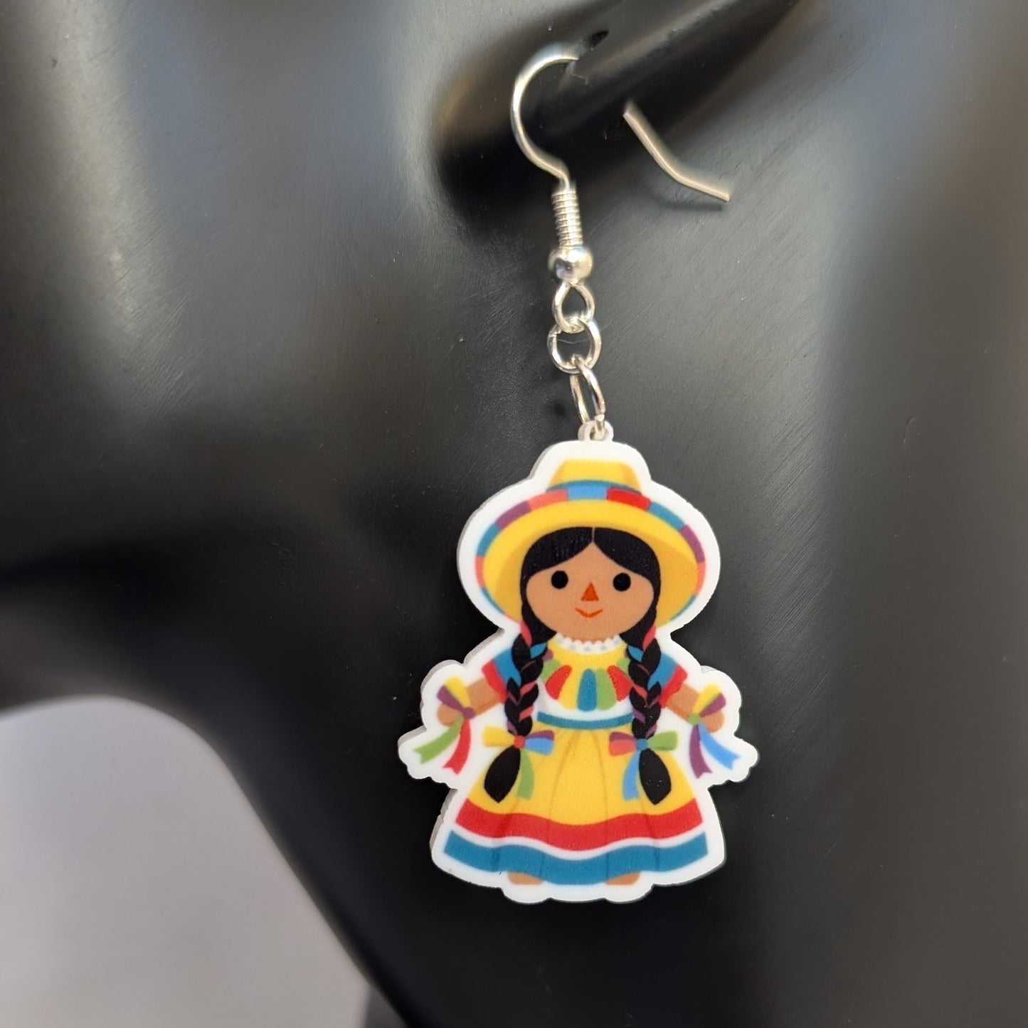 Close-up of a colorful Muñeca Mexican doll acrylic dangle earring wearing a traditional yellow dress and hat.