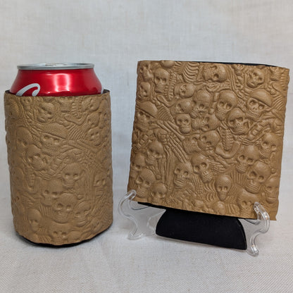 Tan embossed skull pattern leather can cooler holding a red beverage can beside a matching leather sample on a display stand.