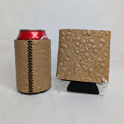 Tan embossed skull pattern leather can cooler with visible side stitching, red beverage can, and matching leather sample on a display stand.