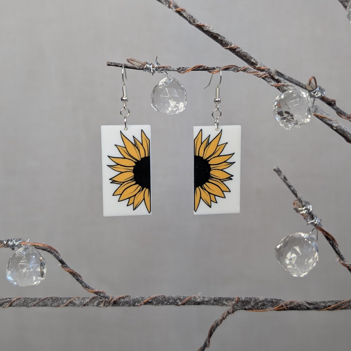 Pair of bright cheerful sunflower dangle earrings with yellow petals, black centers, and white rectangular backgrounds.