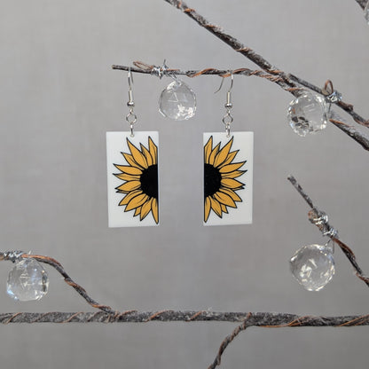 Pair of bright cheerful sunflower dangle earrings with yellow petals, black centers, and white rectangular backgrounds.