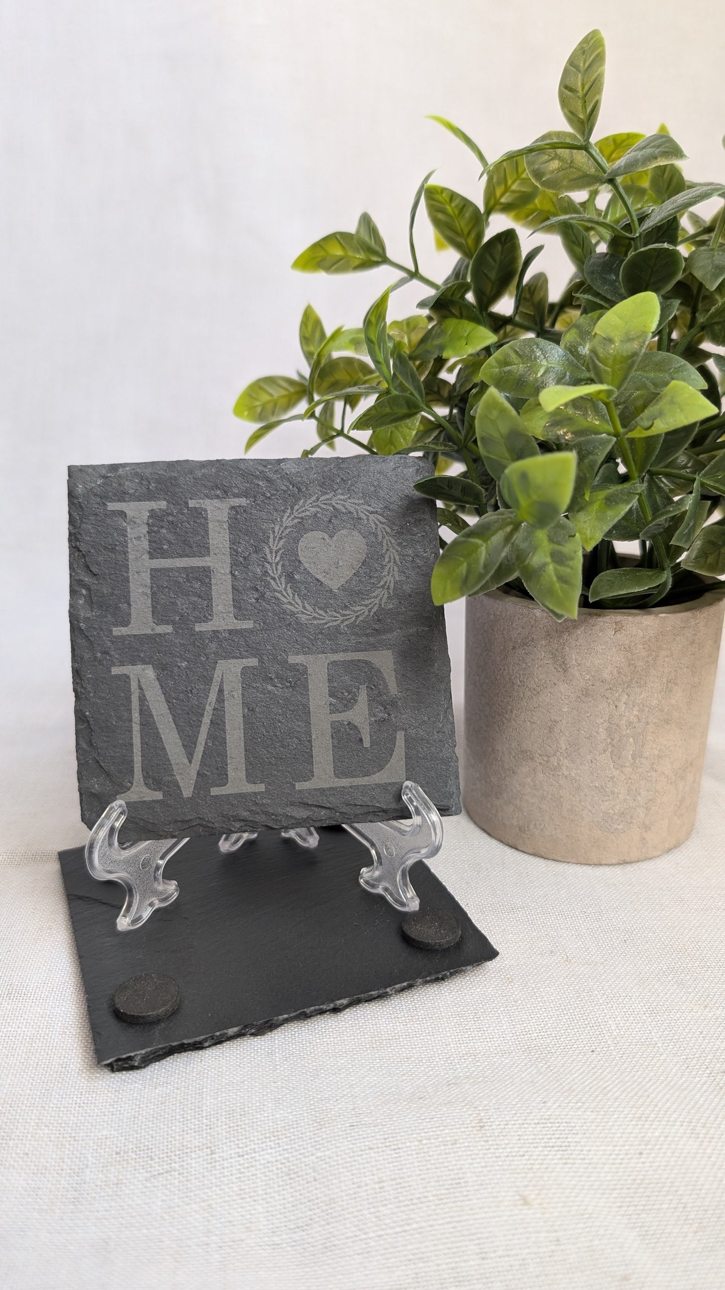 Engraved slate coaster featuring a "HOME" design displayed on a stand beside a decorative plant, ideal for housewarming gifts, realtor closing gifts, and personalized home décor.