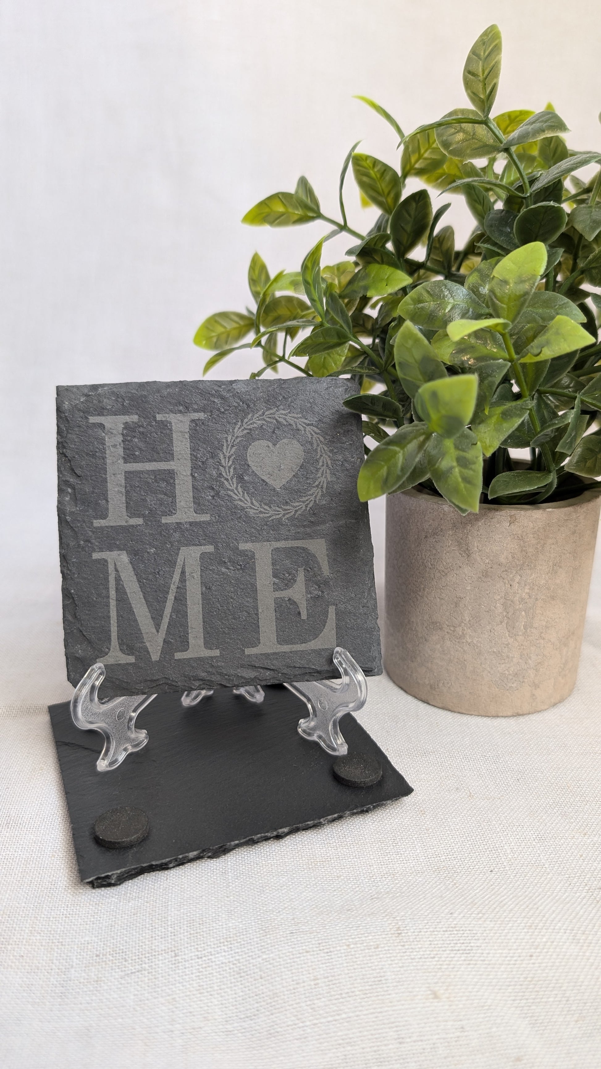 Engraved slate coaster featuring a "HOME" design displayed on a stand beside a decorative plant, ideal for housewarming gifts, realtor closing gifts, and personalized home décor.