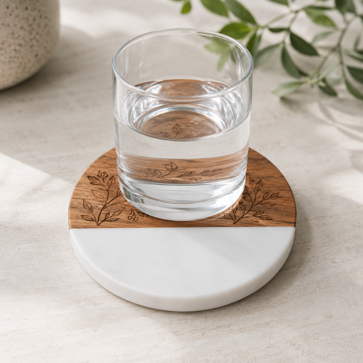 Clear drinking glass resting on a round engraved marble and acacia wood coaster with a botanical design laser engraved into the wood section.