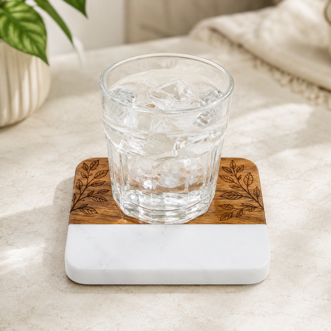 Clear drinking glass resting on a square marble and acacia wood coaster with a botanical design laser engraved into the wood section.