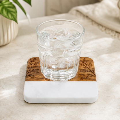 Clear drinking glass resting on a square marble and acacia wood coaster with a botanical design laser engraved into the wood section.