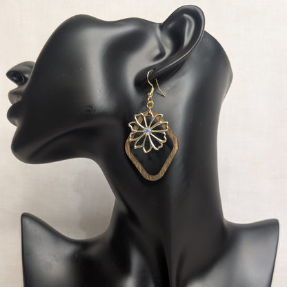 Geometric wood and metal charm earring with a gold floral design and iridescent center stone displayed on a black jewelry stand.