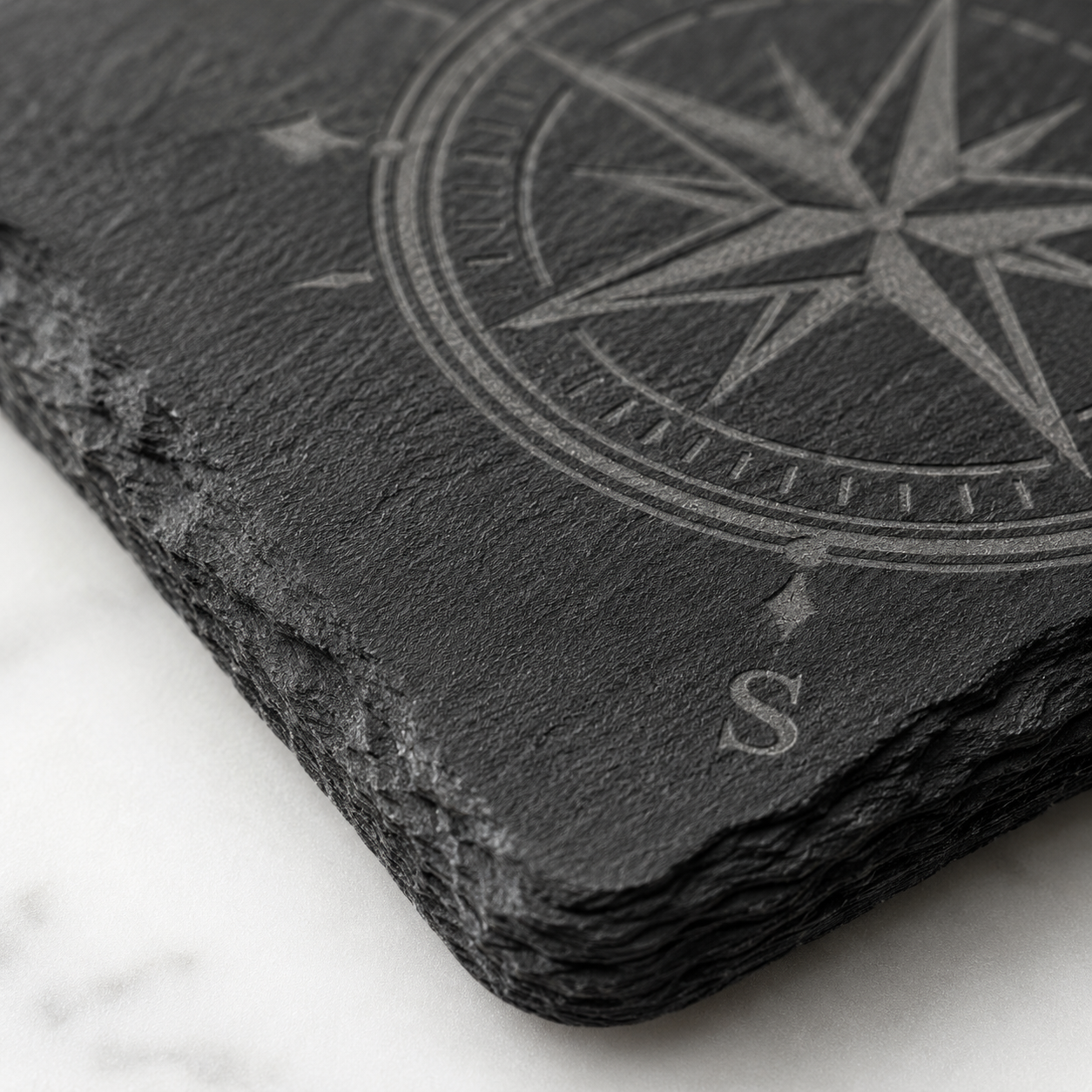 Close-up of a square slate coaster laser engraved with a compass design, showing the natural rough edge and dark stone texture.