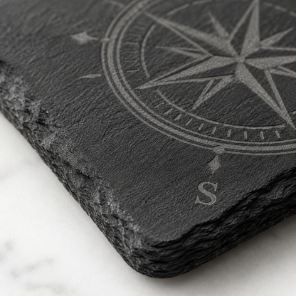 Close-up of a square slate coaster laser engraved with a compass design, showing the natural rough edge and dark stone texture.