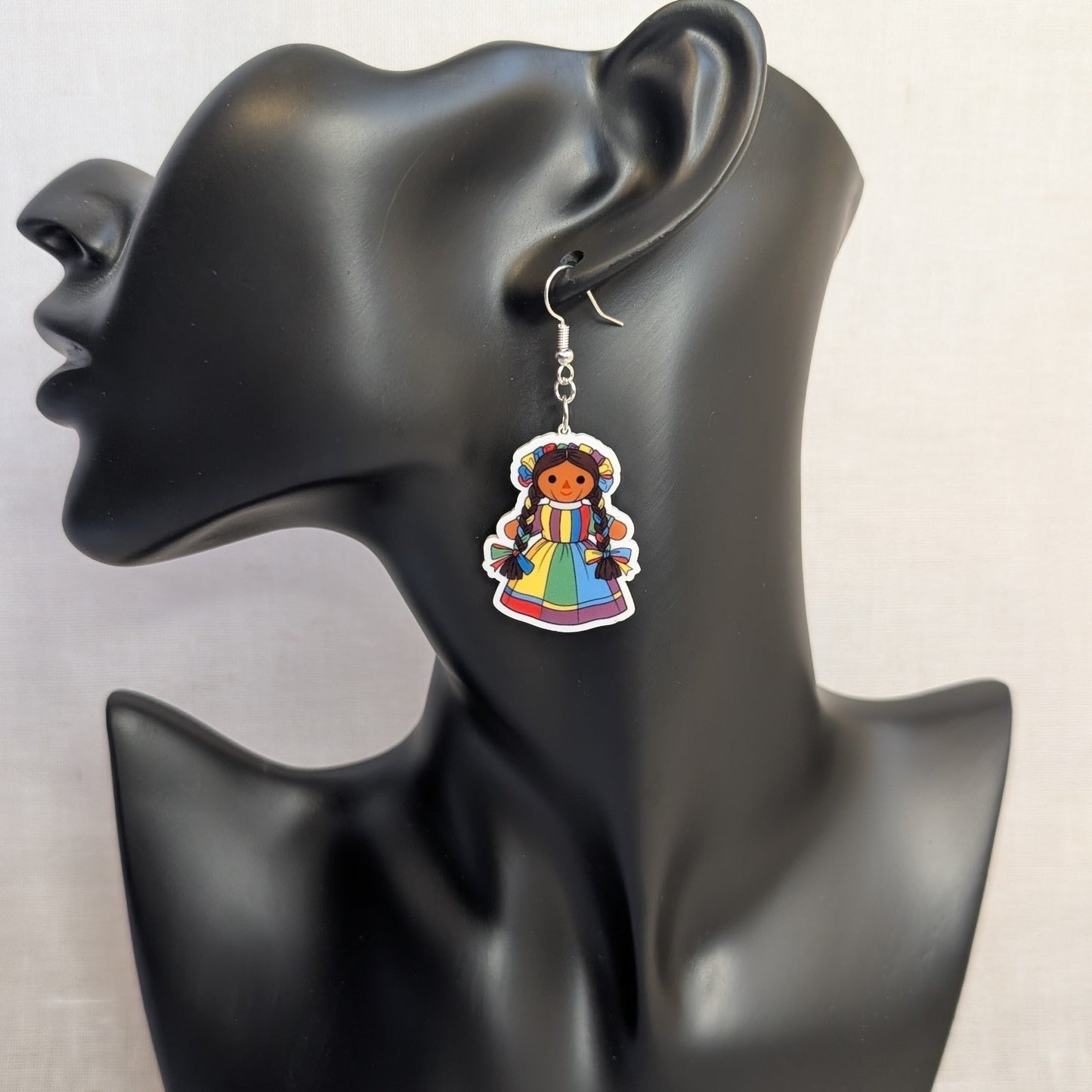 Colorful Muñeca Mexican doll acrylic dangle earring displayed on a black jewelry stand, featuring a traditional multicolor dress and braided hair.
