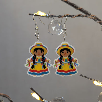 Pair of colorful Muñeca Mexican doll acrylic dangle earrings featuring traditional yellow dresses, braided hair, and matching hats.
