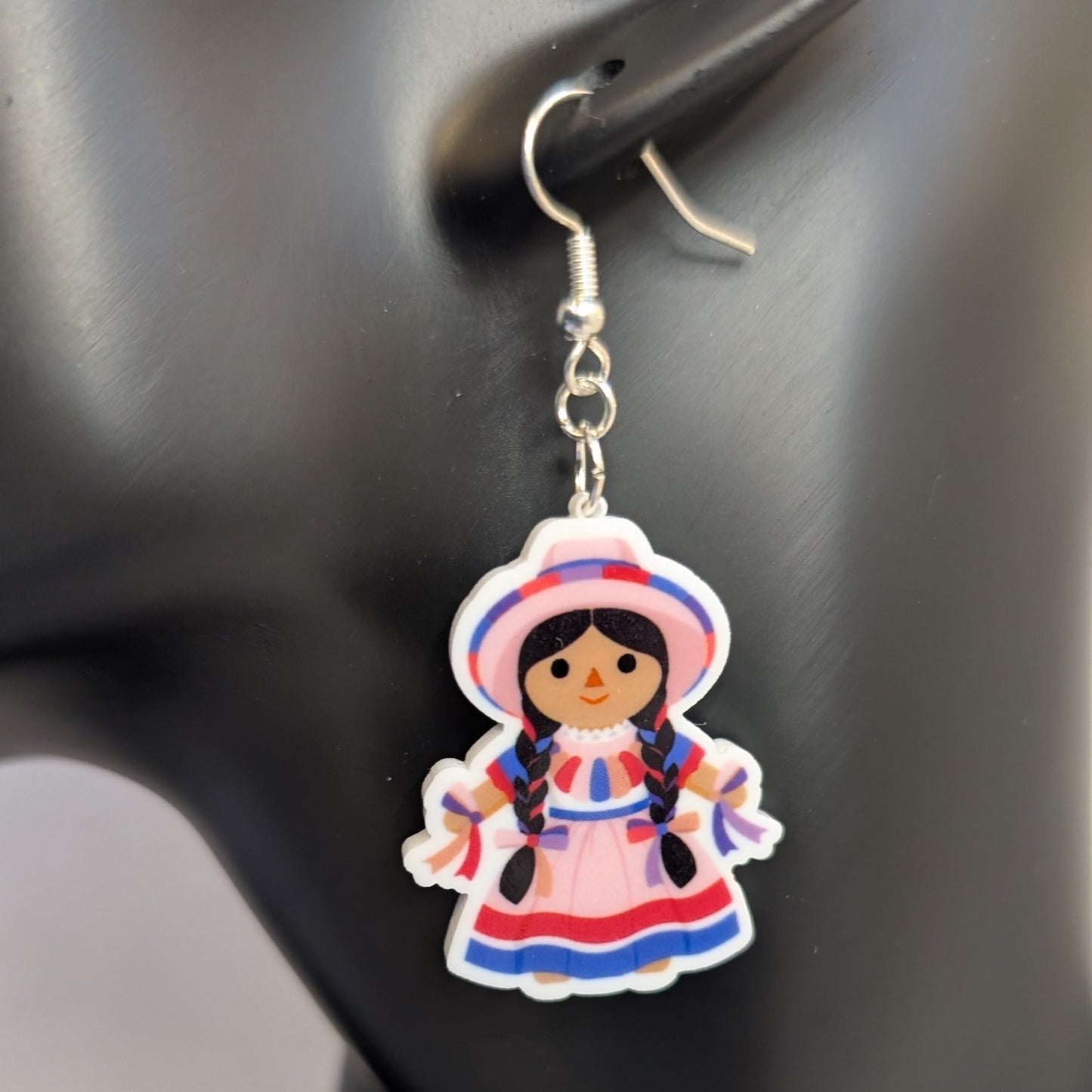 Close-up of a pink Muñeca Mexican doll acrylic dangle earring with braided hair, a traditional dress, and matching hat.