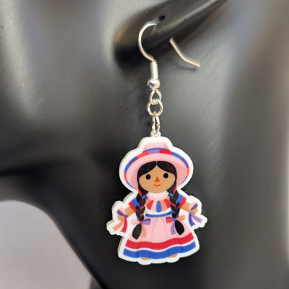Close-up of a pink Muñeca Mexican doll acrylic dangle earring with braided hair, a traditional dress, and matching hat.