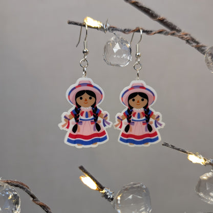 Pair of pink Muñeca Mexican doll acrylic dangle earrings with braided hair, traditional dresses, and matching hats.