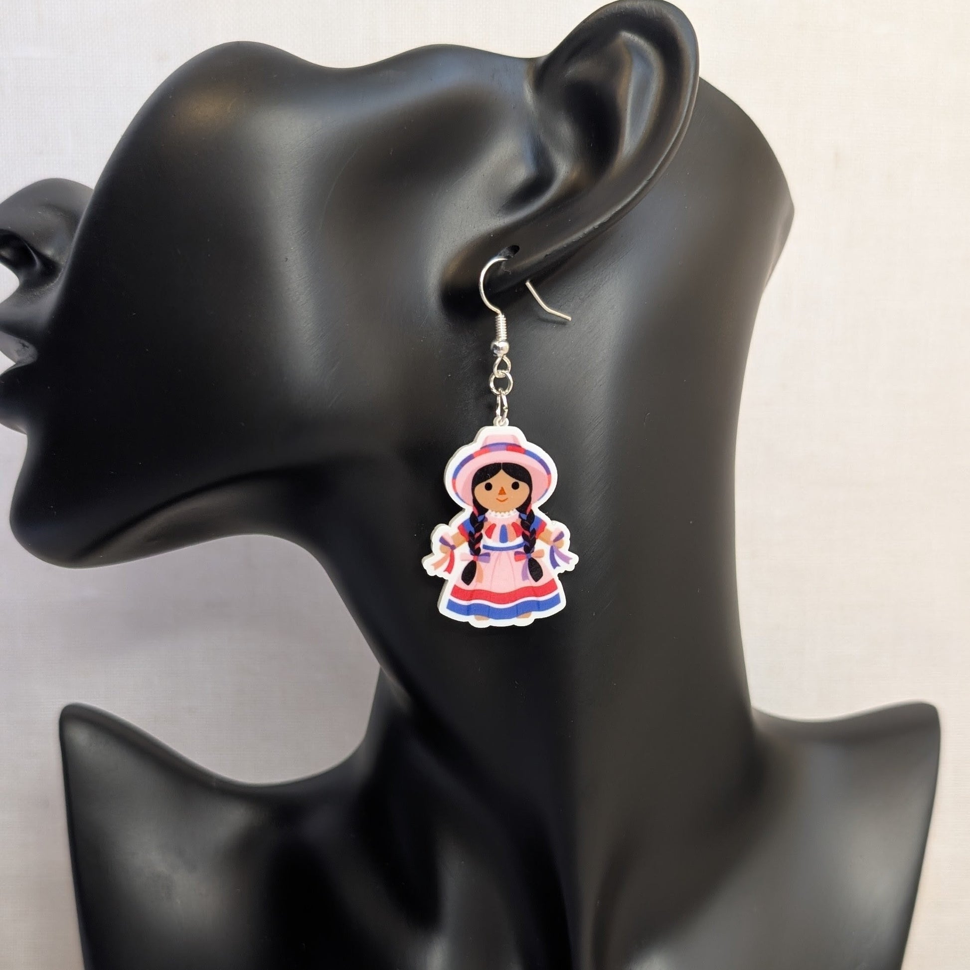 Pink Muñeca Mexican doll acrylic dangle earring displayed on a black jewelry stand, featuring a traditional dress, braided hair, and matching hat.