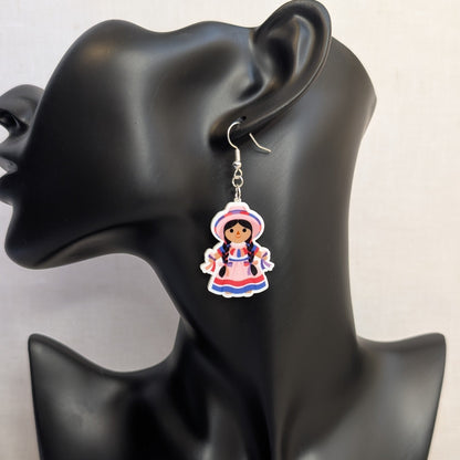 Pink Muñeca Mexican doll acrylic dangle earring displayed on a black jewelry stand, featuring a traditional dress, braided hair, and matching hat.