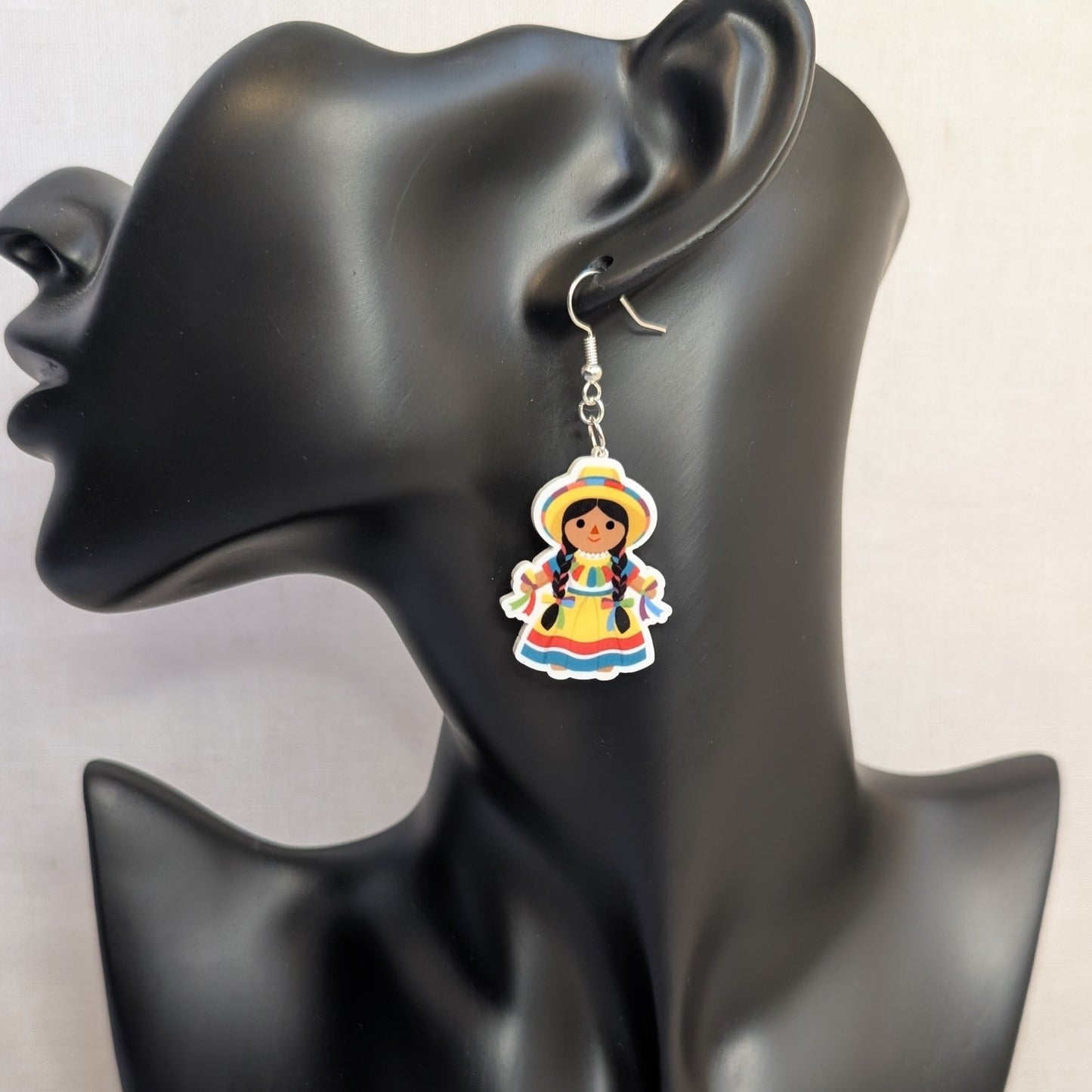 Colorful Muñeca Mexicana doll dangle earring displayed on a black jewelry stand, featuring a traditional Mexican dress and hat.