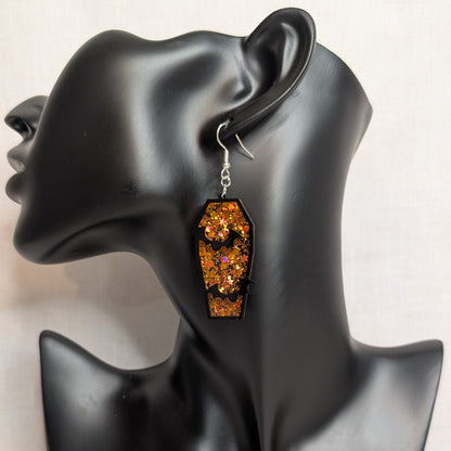 Orange glitter coffin dangle earring with black bat cutouts displayed on a black jewelry mannequin for Halloween.