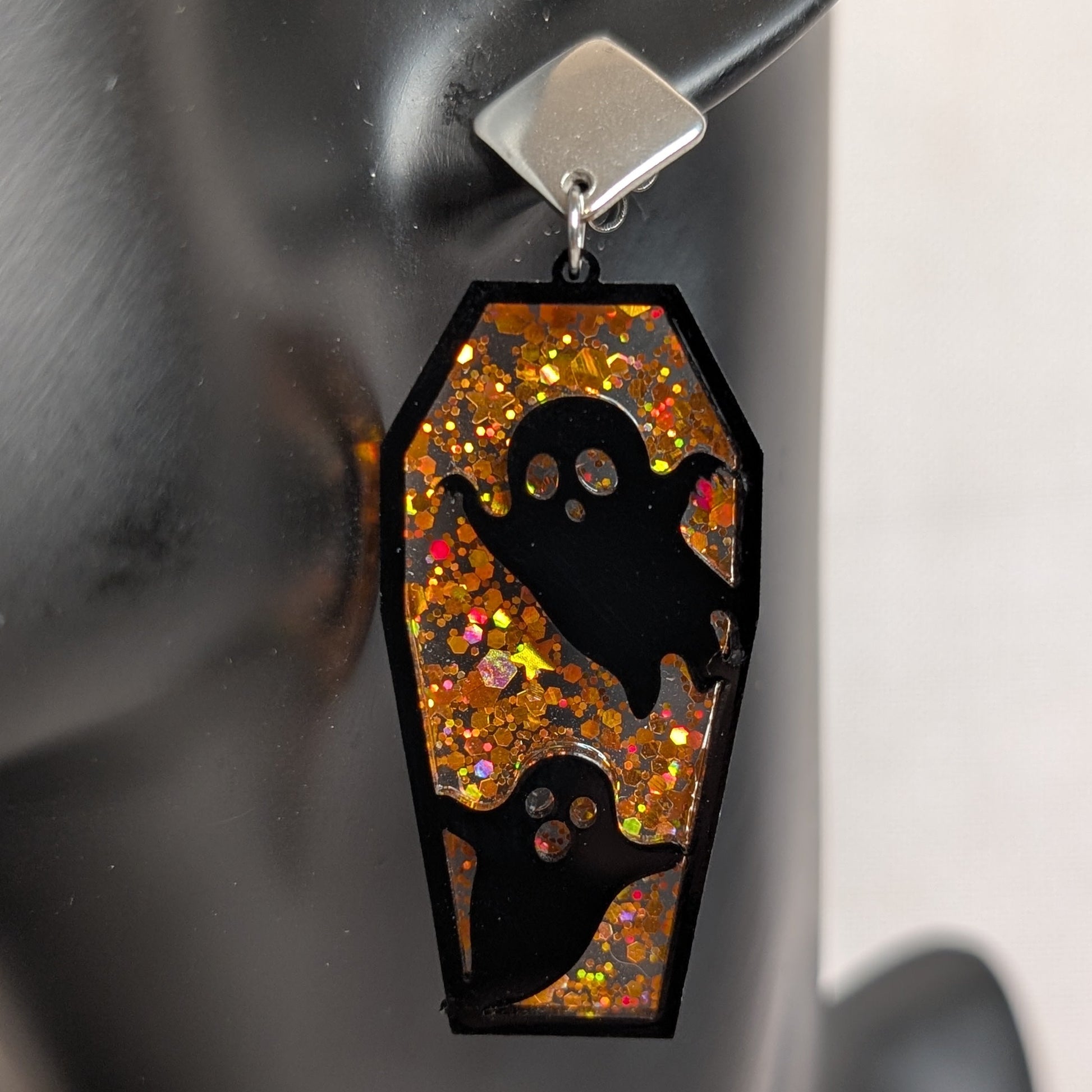 Orange glitter coffin-shaped dangle earring with black ghost cutouts, handmade Halloween statement jewelry.