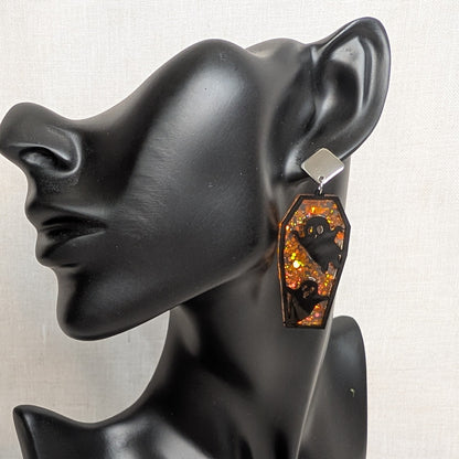 Orange glitter coffin-shaped dangle earring with black ghost cutouts displayed on a black jewelry mannequin.