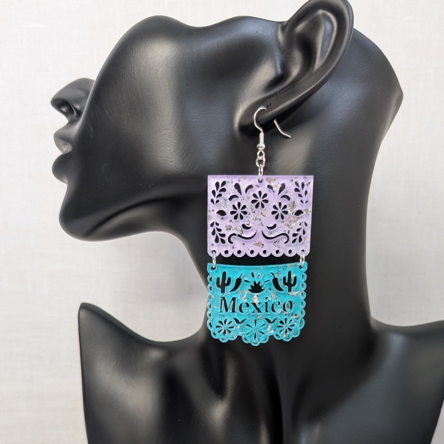 Turquoise and white Papel Picado Mexico dangle earring with floral cutout details displayed on a black jewelry stand.