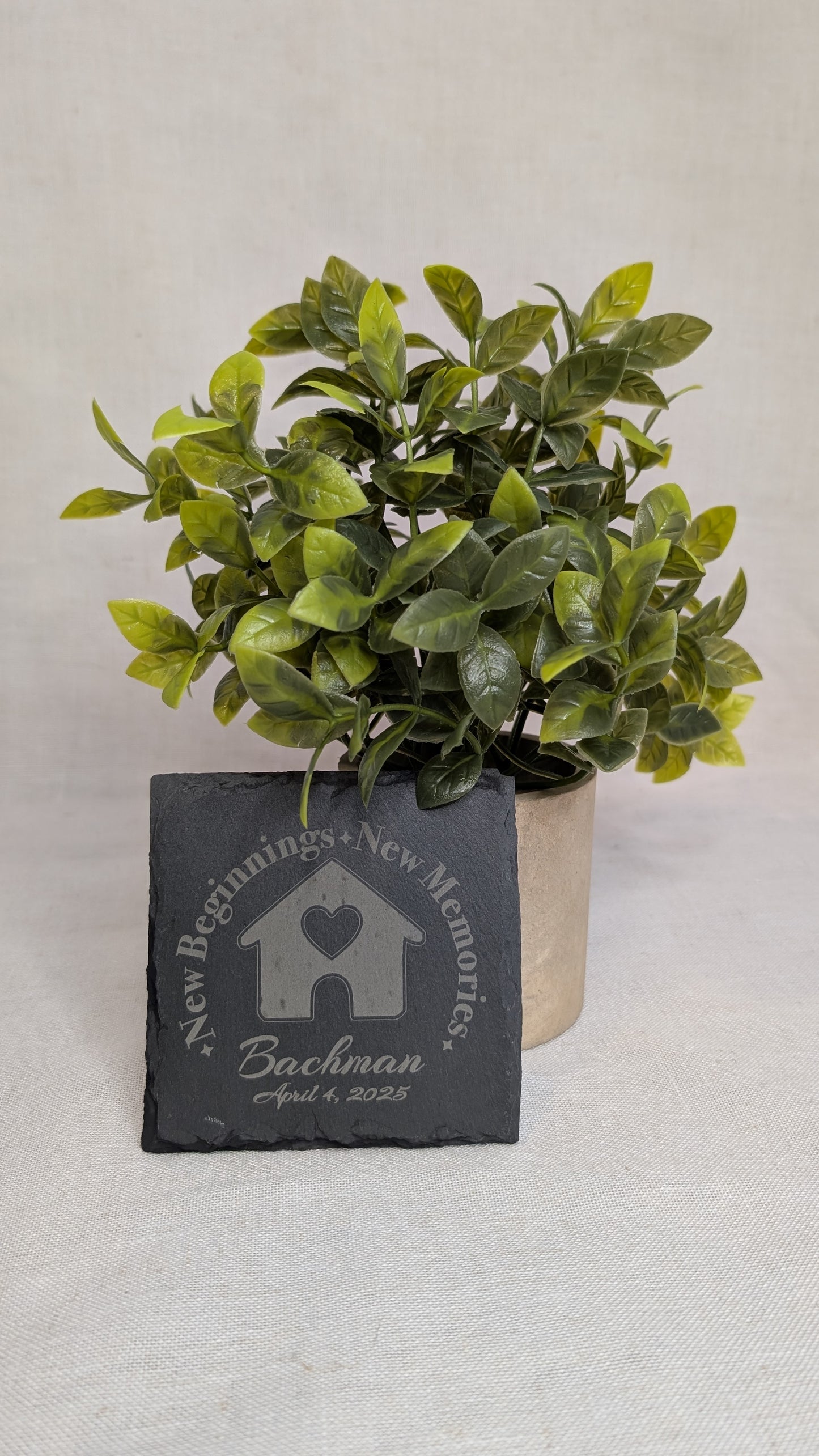 Personalized engraved slate coaster featuring a "New Beginnings, New Memories" home design with custom family name and move-in date, displayed beside a decorative plant. Ideal for realtor closing gifts and housewarming gifts.