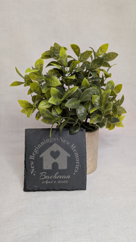 Personalized engraved slate coaster featuring a "New Beginnings, New Memories" home design with custom family name and move-in date, displayed beside a decorative plant. Ideal for realtor closing gifts and housewarming gifts.