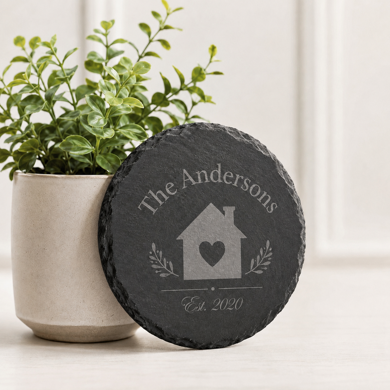 Personalized round slate coaster engraved with a family name, house design, heart, and established date beside a plant.