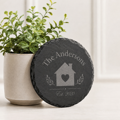 Personalized round slate coaster engraved with a family name, house design, heart, and established date beside a plant.