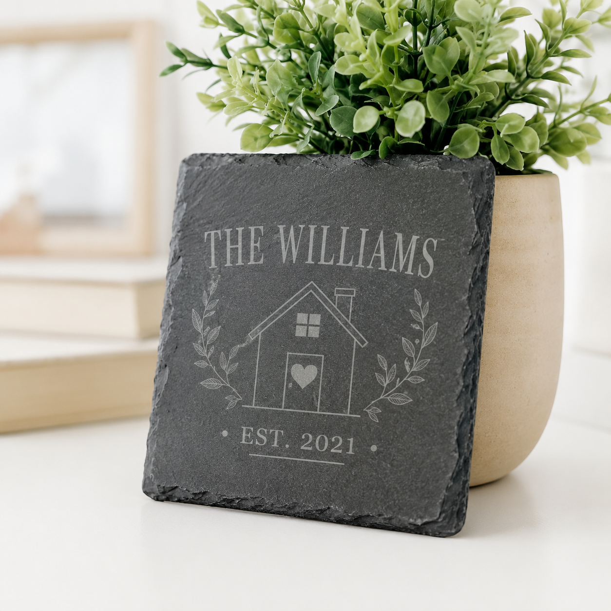 Personalized square slate coaster engraved with the Williams family name, house design, heart, and established date beside a potted plant.