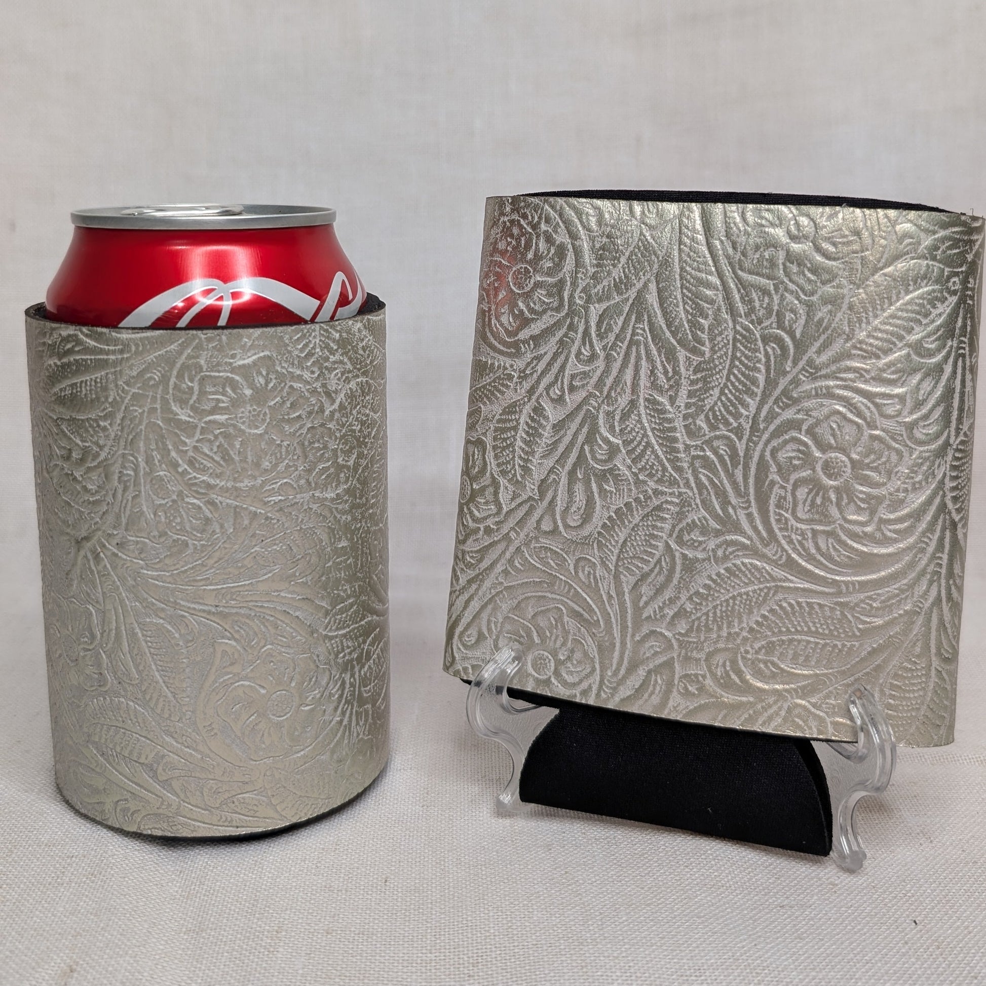 Silver metallic embossed floral leather can cooler holding a red beverage can beside a matching floral leather sample on a display stand.