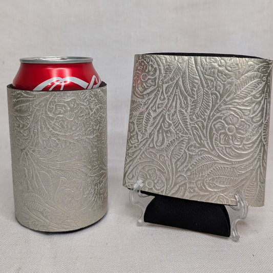 Silver metallic embossed floral leather can cooler holding a red beverage can beside a matching floral leather sample on a display stand.