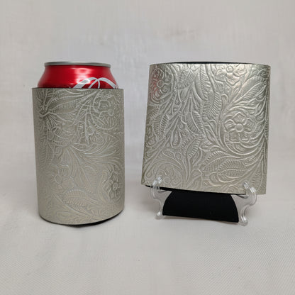Silver metallic embossed floral leather can cooler holding a red beverage can beside a matching embossed leather sample on a display stand.