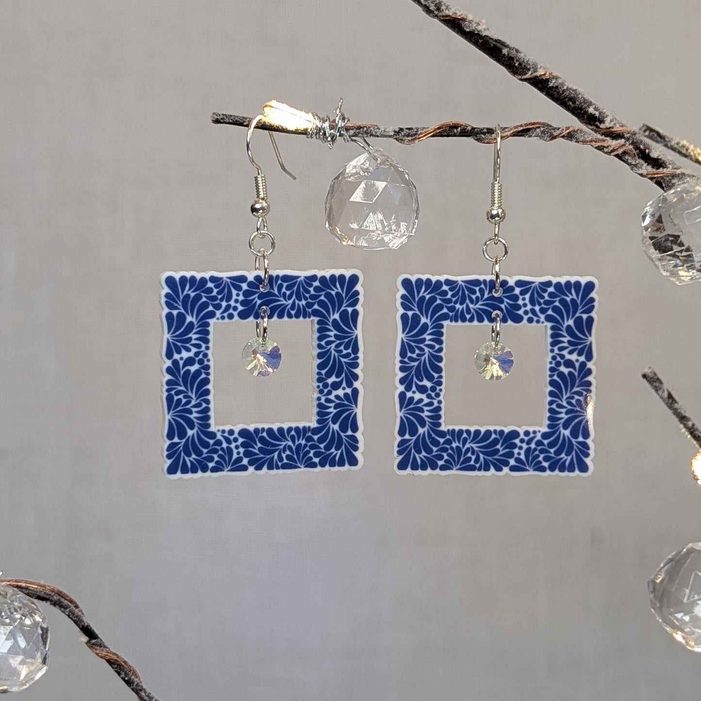 Blue Talavera-style picture frame earrings with white floral patterns and iridescent crystal dangle charms.