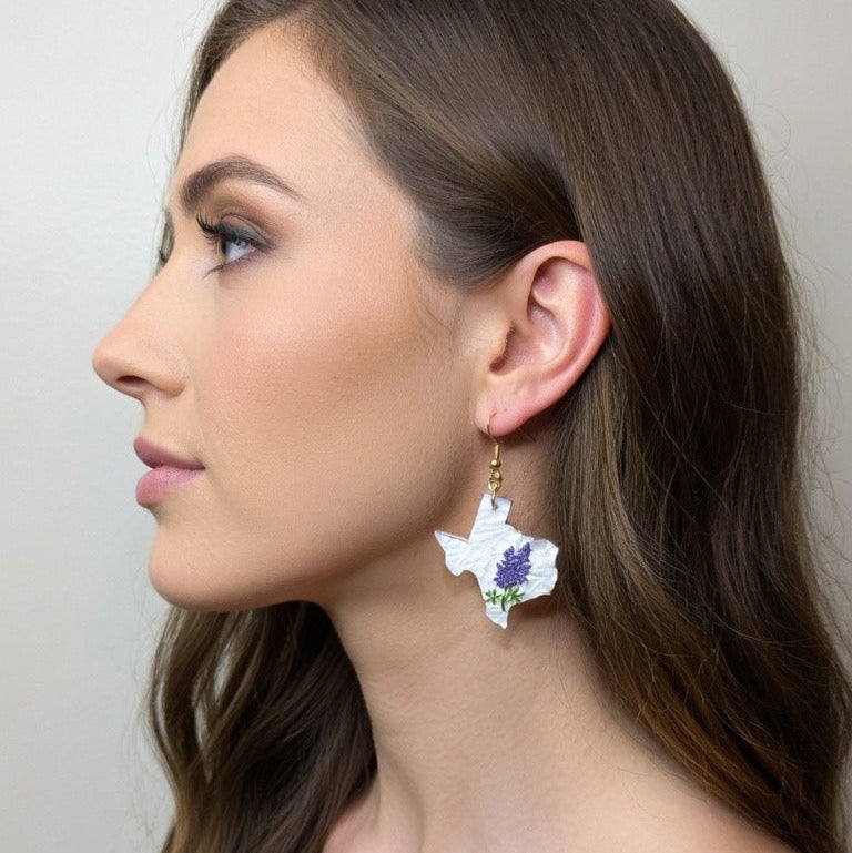 Woman wearing a white Texas-shaped dangle earring featuring bluebonnet flowers and a gold hook.