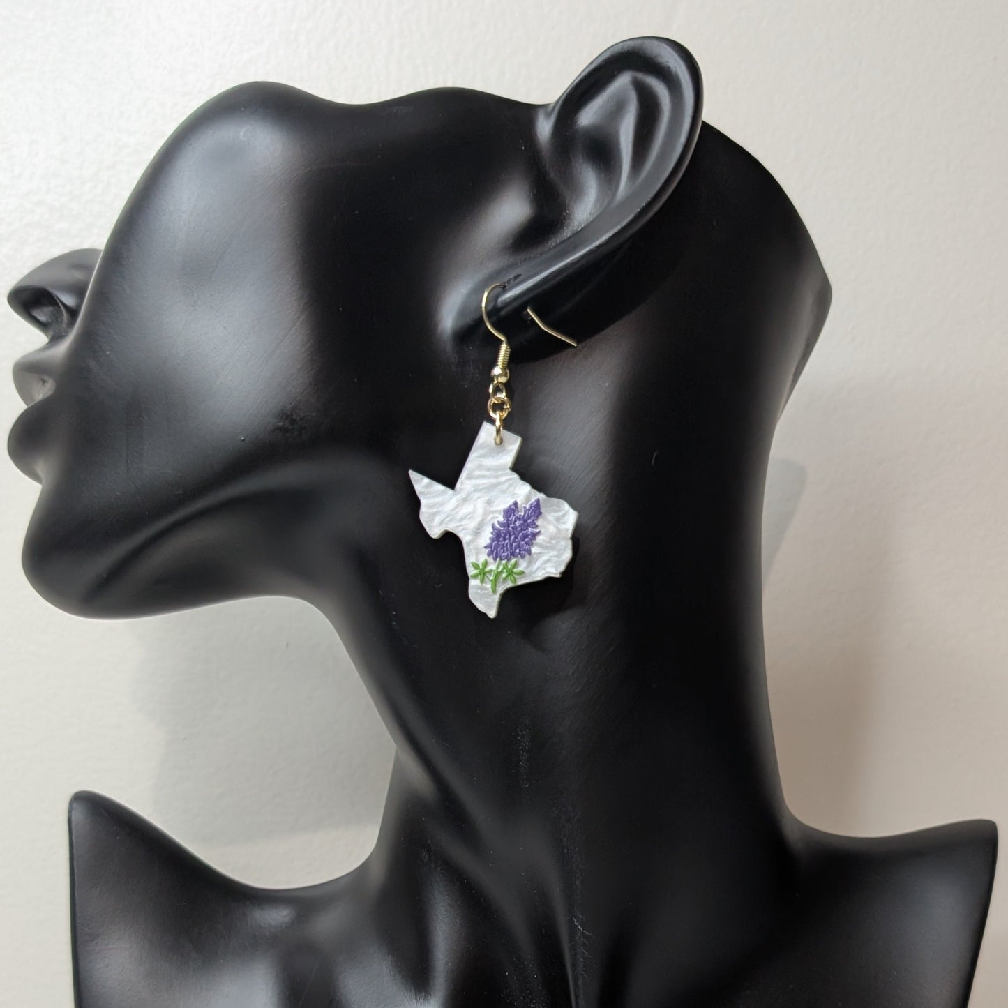 White Texas-shaped dangle earring with bluebonnet flowers displayed on a black jewelry stand.