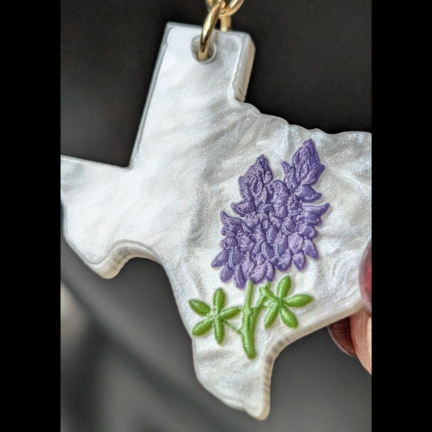 Macro close-up of a white Texas-shaped dangle earring featuring detailed bluebonnet flowers, green leaves, and a gold hook.