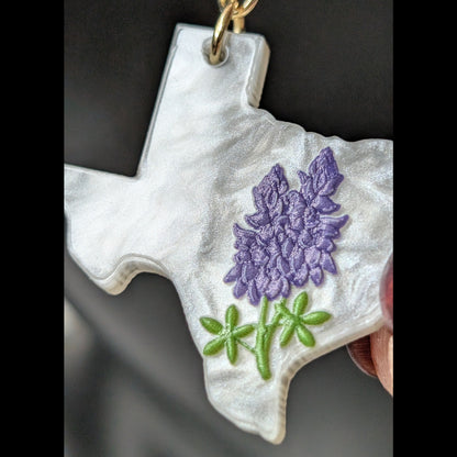 Macro close-up of a white Texas-shaped dangle earring featuring detailed bluebonnet flowers, green leaves, and a gold hook.